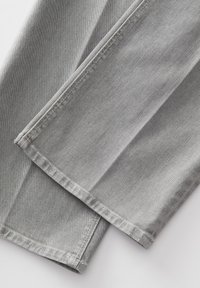 Lichtgrijze denim broekspijpen die over elkaar zijn gevouwen met zichtbare stiksels en textuur op een witte achtergrond.