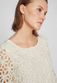 Hvit stofftopp med blomsterlandi teksturdesign, med vid halsringning og åpne skulderpartier. Myk, lagdelt tekstur gjennom hele toppen.