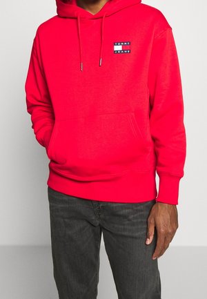 Mies pukeutuneena kirkkaan punaiseen Tommy Jeans -huppariin, jossa on etutasku, ja mustat farkut, seisoo yksiväristä vaaleaa taustaa vasten.