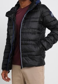 Czarna puchowa kurtka z kapturem, z pikowanym wzorem, niebieskimi zamkami i kieszeniami bocznymi. Nosi się ją na bordowej koszulce.