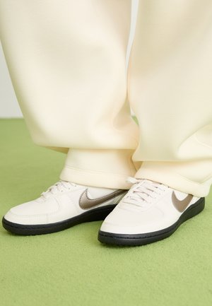 Baskets blanches en cuir avec un swoosh gris, semelle en caoutchouc noir et lacets blancs, portées avec un jogging crème sur une surface verte.