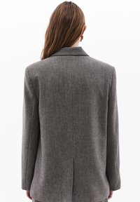 Harmaa tweed-sakko, jossa on rakenteellinen muotoilu, leikattu kaulus ja keskitakamus. Varustettu pitkillä hihoilla ja teksturoidulla kankaalla.