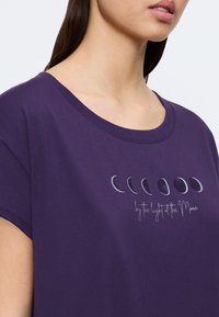 Fioletowa bawełniana koszulka z krótkim rękawem, z nadrukiem faz księżyca i napisem "przy blasku Księżyca" poniżej.