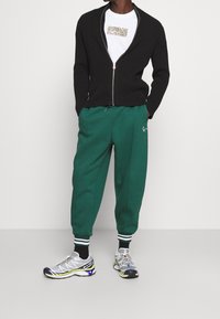 Černý žebrovaný svetr na zip, bílý tričko s grafikou, zelené zúžené tepláky a stříbrné sportovní boty s modrými a žlutými akcenty.