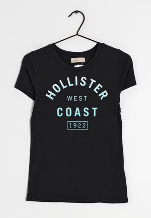 Camiseta negra de manga corta con texto en azul claro que dice "HOLLISTER WEST COAST 1922" en un diseño audaz y curvado. Exhibida en una percha.