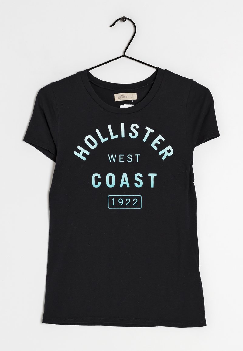 Sort t-shirt med korte ærmer og lyseblå tekst, der lyder "HOLLISTER WEST COAST 1922" i et fedt, buet design. Vist på en bøjle.