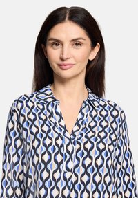 Blouse à motifs en tissu léger présentant un motif ondulé répétitif bleu et crème. Elle possède un col et une fermeture à boutons sur le devant.