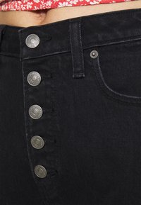 Black denim jeans with a high-rise waist, featuring five metallic buttons on the front, visible stitching, and a textured fabric.