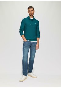 Felpa verde acqua con collo a cowl e lacci, abbinata a jeans blu chiaro scoloriti e sneakers bianche, con una finitura testurizzata.
