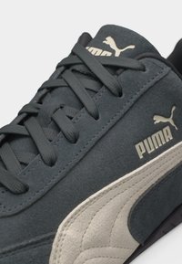 Σκούρο γκρι σουέντ αθλητικό παπούτσι Puma με μπεζ πλευρική ρίγα, μαύρα κορδόνια και λογότυπο Puma στη γλώσσα και στην πλευρά.
