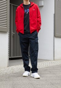 Rode hoodie met rits en kangoeroezakken over een zwart T-shirt, gecombineerd met marineblauwe cargobroek met meerdere zakken en witte sneakers.