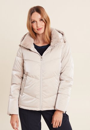 Pufferjack in lichtbeige met een imitatiebonten capuchon. Heeft diagonale stiksels, een ritssluiting aan de voorkant en lange mouwen. Draagt over een donkere top.