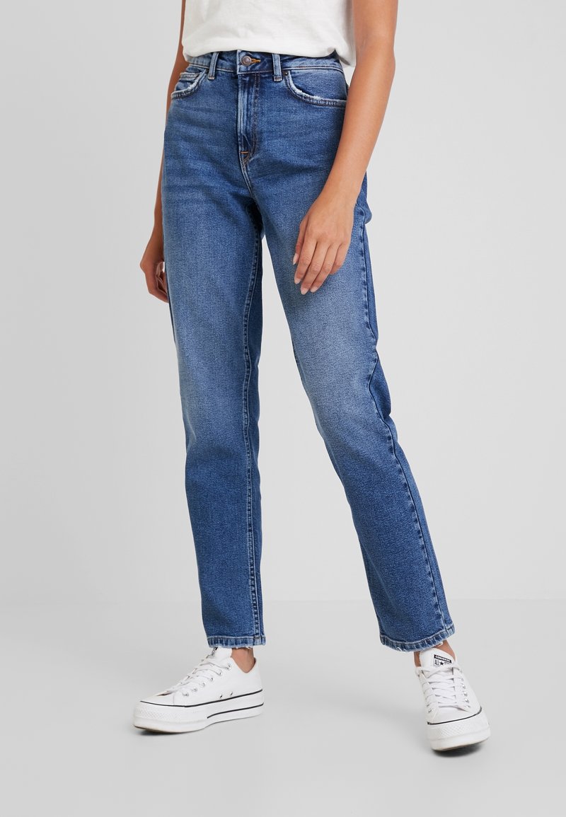 Jean en denim bleu taille haute avec une coupe droite, une légère décoloration et des coutures contrastées. Associé à des baskets en toile blanches.