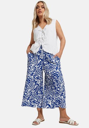 Witte linnen top met een vetersluiting aan de voorkant, gecombineerd met blauwe en witte, gebloemde wijde broeken met zijzakken. Witte platte sandalen.