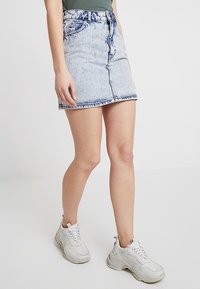 Osoba v světle modré mini sukni z acid wash jeanu, zeleném ribbed topu a bílých chunky teniskách stojící proti jednoduchému pozadí.