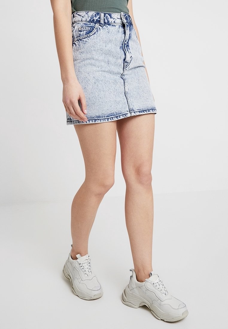 Osoba v světle modré mini sukni z acid wash jeanu, zeleném ribbed topu a bílých chunky teniskách stojící proti jednoduchému pozadí.