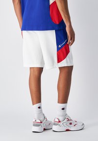 Wei�ße Shorts mit roten und blauen Akzenten, kombiniert mit weißen Sneakern und gemusterten Socken. Mit einem Logo auf den Shorts und den Sneakern.