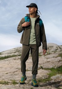 Utendørs antrekk med en brun vanntett jakke, grønn t-skjorte, brune bukser og flerfargede tursko, supplert med en teal ryggsekk.
