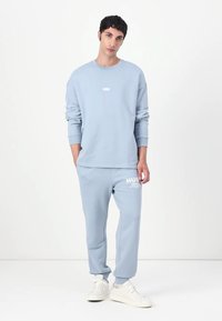 Világoskék pulóver és melegítőnadrág puha pamutszövetből. A pulóver kis logóval díszített, a nadrágon fehér grafikai nyomtatás található. Az outfitt fő eleme egy pár sportcipő.