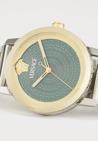 Reloj dorado con una esfera teal de patrón, que presenta un emblema de león, manecillas doradas y una correa de acero inoxidable con acentos geométricos.