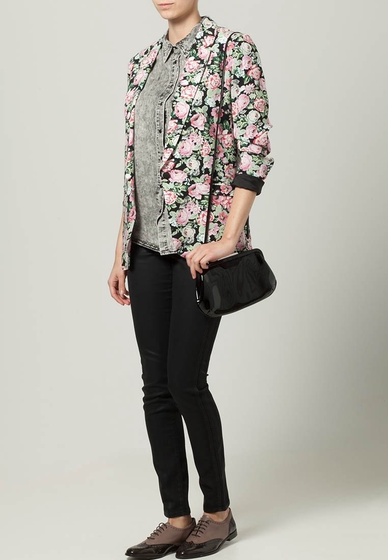 Femme portant un blazer fleuri sur une chemise grise, pantalon skinny noir, chaussures oxford marron, tenant un petit sac noir.