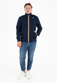 Felpa zip con collo alto di colore blu navy, con una cerniera a strisce multicolori, polsini a coste e tasche laterali. Indossata con jeans chiari.