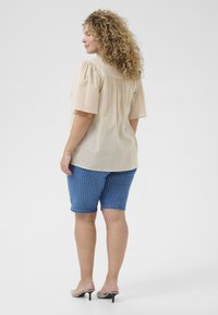 Beige short-sleeve top with gathered shoulders, paired with blue striped denim shorts. Model wears neutral high-heeled sandals.