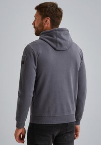 Sudadera con capucha gris fabricada con material suave, que presenta un diseño sencillo, puños acanalados y un bolsillo de parche en la manga izquierda.