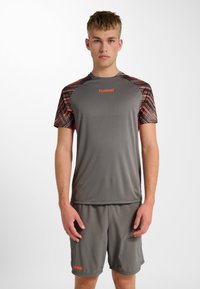 Jonge man die grijze sportkleding met oranje accenten en "hummel"-logo's draagt, staand tegen een effen witte achtergrond, voorover gericht.