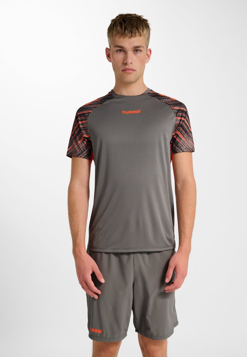 Jonge man die grijze sportkleding met oranje accenten en "hummel"-logo's draagt, staand tegen een effen witte achtergrond, voorover gericht.
