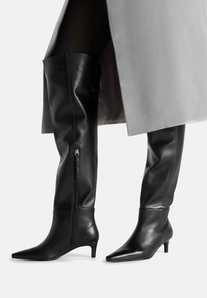 Czarne skórzane kozaki do kolan z szpiczastymi noskami, wyposażone w boczny zamek błyskawiczny i niski obcas typu kiciuś, na szarym tle.