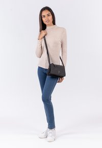 Schwarze Umhängetasche mit strukturierter Oberfläche, Reißverschluss und Logodetail. Modell trägt ein beiges Oberteil, blaue Jeans und weiße Sneakers.