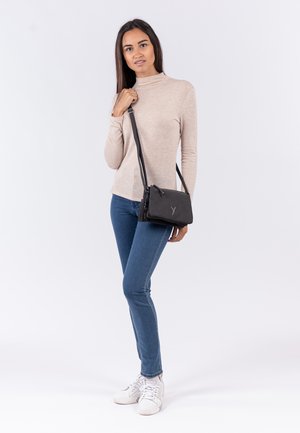Black crossbody bag with textured surface, zip closure, and logo detail. Model wears beige top, blue jeans, and white sneakers.