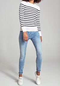 Donna che indossa un top a maniche lunghe bianco e blu a righe, jeans skinny azzurri chiari e sneakers bianche, in piedi su uno sfondo neutro.