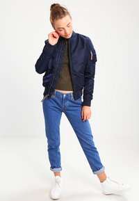 Chaqueta de bombardero azul marino con textura suave, puños y cuello acanalados; combinada con jeans de mezclilla azul claro arremangados y zapatillas blancas.
