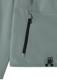 Gros plan sur la poche verticale zippée noire d'une veste verte et le cordon de serrage réglable à l'ourlet.