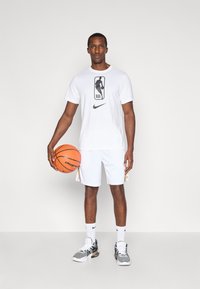 Balts Nike T-krekls ar NBA logo, balti šorti, melni un pelēki sporta apavi, tur rokās oranžu basketbola bumbu. Vienkāršs fons.