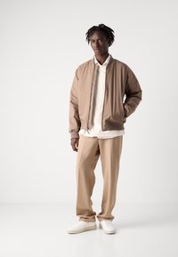 Tan bomberjack met een crèmekleurige sweatshirt, gecombineerd met tan broeken en witte sneakers. Eenvoudig ontwerp, met ribbels aan de mouwen en zijzakken.