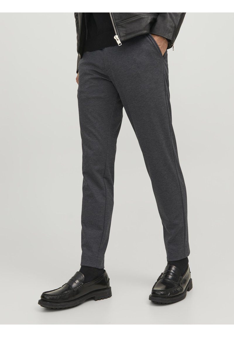 Homme portant un pantalon gris foncé ajusté, des chaussettes noires, des mocassins en cuir noir, et une veste noire partiellement visible avec une fermeture éclair.
