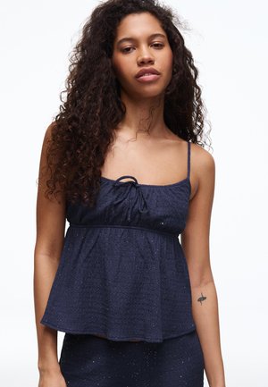 Nő hosszú, göndör hajjal, sötétkék ujjatlan felsőt visel, amelynek vékony pántjai és a nyakkivágásnál egy kis masni található, fehér háttér előtt.