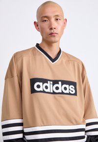 Άντρας με ξυρισμένο κεφάλι φοράει ανοιχτόχρωμο αθλητικό μπλουζάκι Adidas με πλέγμα και μαύρες και άσπρες ρίγες στις μανσέτες, στέκεται μπροστά σε απλό ανοιχτό φόντο.