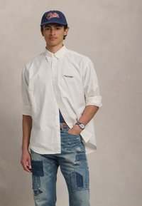 Cămașă albă cu nasturi și logo, blugi denim blue cu detalii de patchwork și uzură. Capac bleumarin și ceas de mână cu cadran rotund.