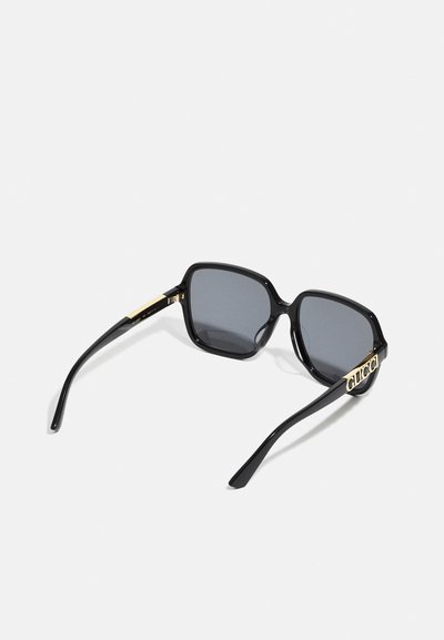 Lunettes de soleil noires carrées avec des lentilles grises. Présentent des montures brillantes, des accents dorés sur les branches et un design moderne.