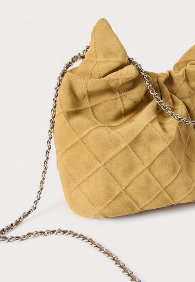 Sac à main en daim matelassé jaune moutarde doux avec une chaîne argentée sur un fond clair uni.