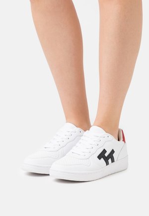 Bílé tenisky s hladkým koženým povrchem, černým logem, perforovaným vzorem na špičce a červeným akcentem na patě.