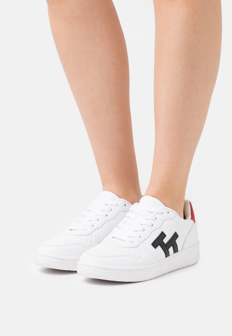 Białe sneakersy z gładką skórzaną powierzchnią, z czarnym logo, perforowanym wzorem na palcach i czerwonym akcentem na pięcie.