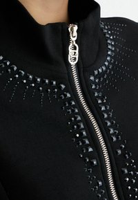 Giacca nera con zip, caratterizzata da borchie decorative nere sulle spalle e sul petto. La cerniera è argentata, a esaltare il design complessivo.