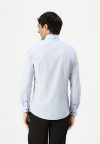 Chemise à manches longues bleu clair avec ourlet arrondi, empiècement au dos et poignets boutonnés. Tissu lisse avec une texture subtile.