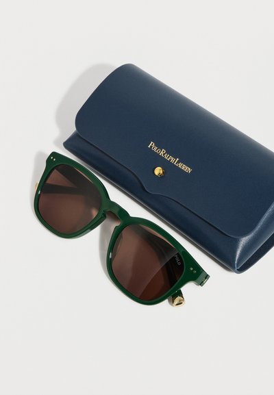 Σκούρα πράσινα γυαλιά ηλίου με καφέ φακούς δίπλα σε μια κλειστή θήκη Polo Ralph Lauren σε ναυτικό μπλε με χρυσά γράμματα και κούμπωμα.