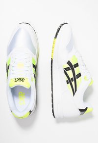 Scarpe sportive bianche con dettagli in argento e giallo neon, caratterizzate da una suola nera testurizzata e un design a strisce nere sui lati.